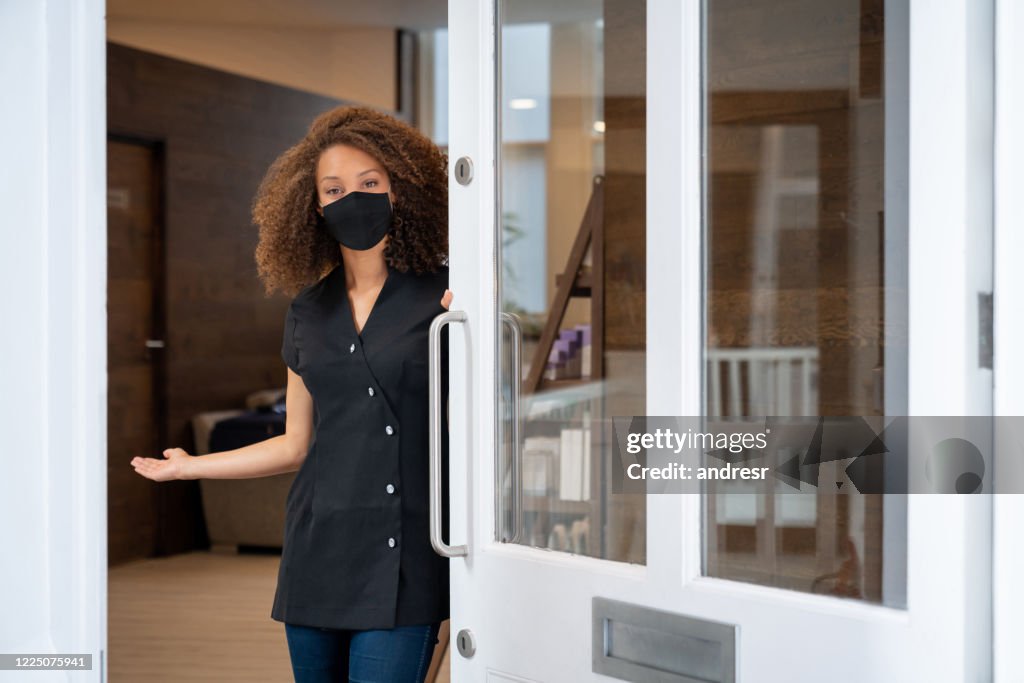 Business owner opening the door of her spa wearing a facemask