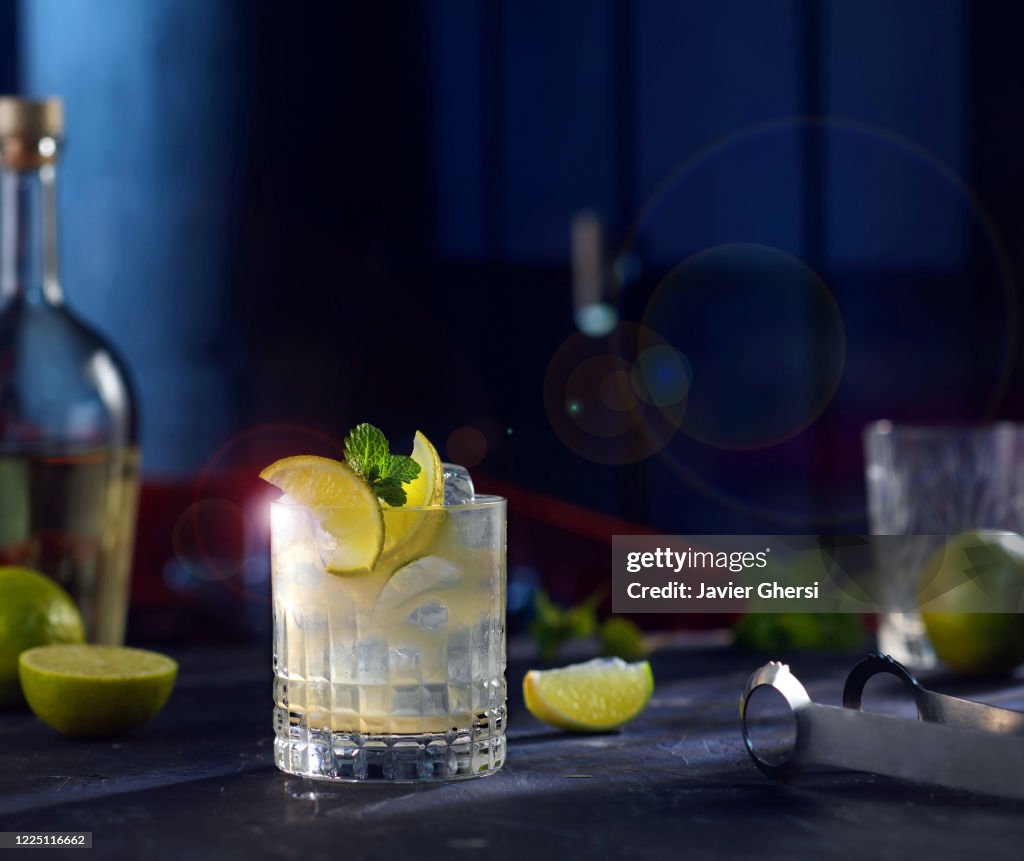 Vaso de whisky sour, rodajas de limón y botellas de fondo