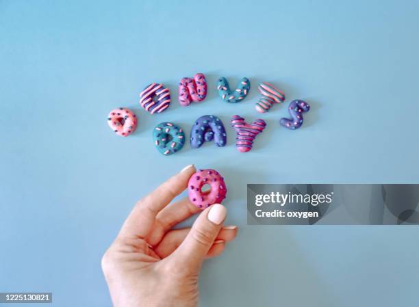 colorful donuts day letters text on blue background made with plasticine. woman hand holding letter. national donut day - human finger print stock pictures, royalty-free photos & images
