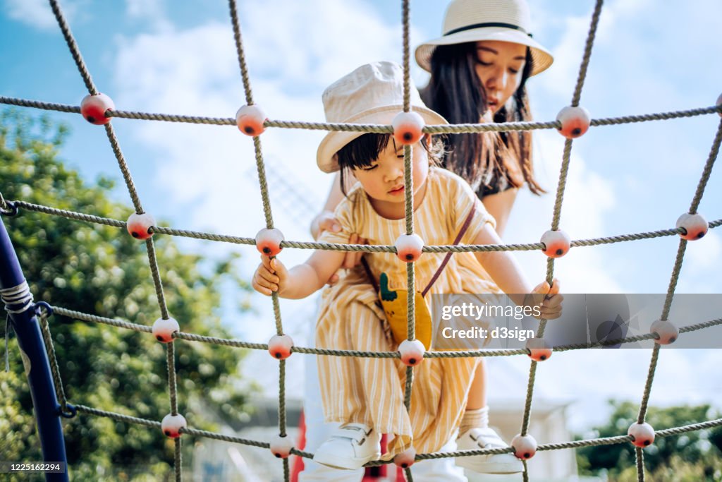 Young Asian mother assisting little daughter on climbing ropes in an outdoor playground on a sunny day