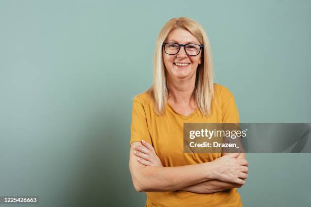 mature blond woman with blue eyes in yellow t-shirt against green or gray wall - woman 50s blue eyes stock pictures, royalty-free photos & images