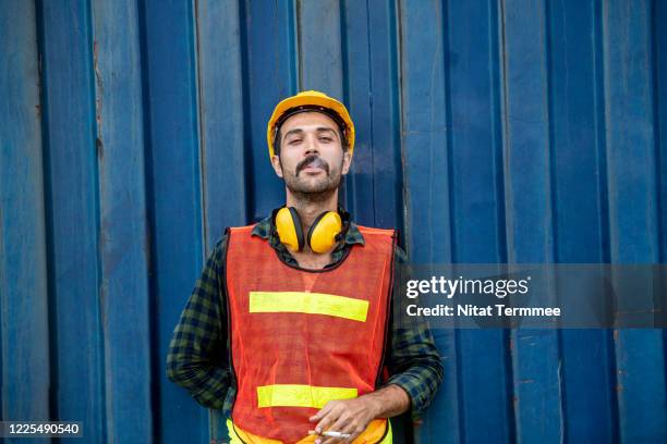 portrait of a bearded worker taking a break and smoking a cigarette while leaning on front of cargo container, wearing hard hat and safety protect suit. - i quit stock pictures, royalty-free photos & images