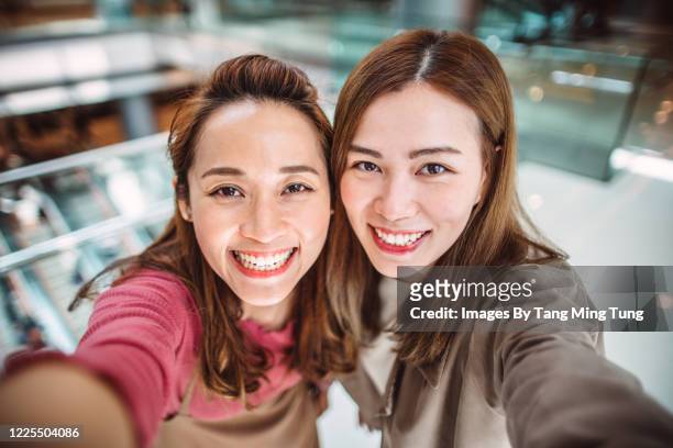 two pretty girlfriends taking selfies with big smiles in shopping mall. - chinese young adults shopping imagens e fotografias de stock