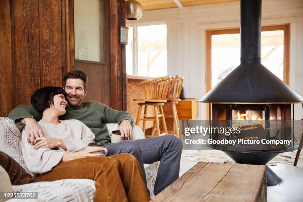 pareja joven riendo bebiendo té y relajándose junto a un fuego en su cabaña - fireplace fotografías e imágenes de stock