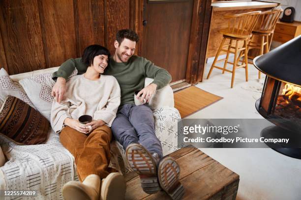 couple talking and laughing while warming up with tea by their fireplace - log cabin fire stock pictures, royalty-free photos & images