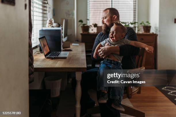 dad working from home with one year old boy crying in his lap - woedeaanval stockfoto's en -beelden