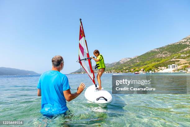 kleines mädchen, das an einem sonnigen tag an einer bucht seinen ersten windsurfunterricht mit einem instruktor nimmt - stock photo - windsurfbrett stock-fotos und bilder