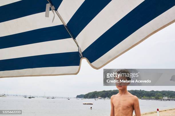 Young boy with brown curly hair brown eyes looking away from the camera striped umbrella in the top of the image boy looking into the distance shirtless water in the background horizon over the water sand and beach in the back of the photo