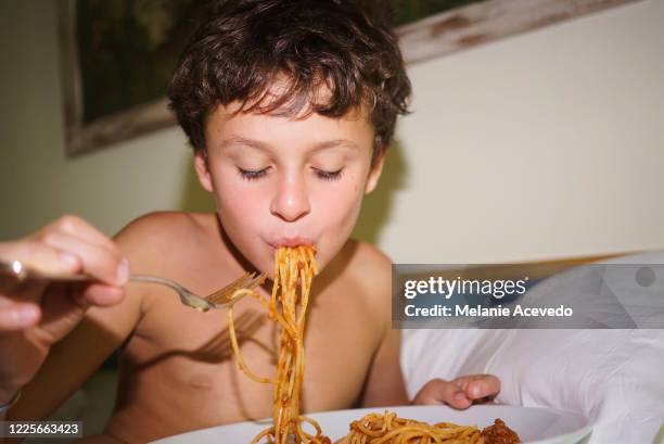 young boy with short brown curly hair and brown eyes close up camera flash looking down eating spaghetti with. a fork in bed eyes closed shirtless white pillows in the background - camera flashes foto e immagini stock