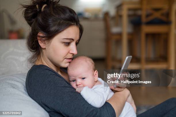 loving and affectionate mother holding newborn baby indoors at home, using smartphone. - new user stockfoto's en -beelden