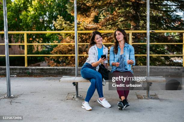 female friends using phone while waiting bus - bus bench stock pictures, royalty-free photos & images