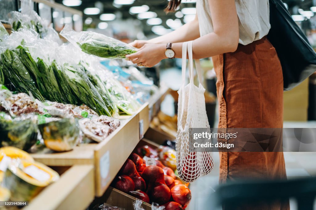 Cropped shot of young Asian woman shopping for fresh organic groceries in supermarket. She is shopping with a cotton mesh eco bag and carries a variety of fruits and vegetables. Zero waste concept