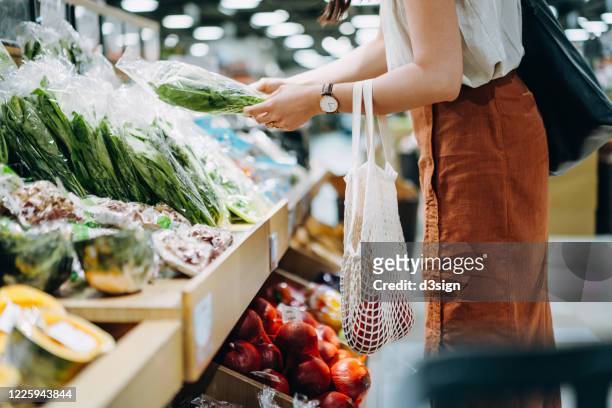 cropped shot of young asian woman shopping for fresh organic groceries in supermarket. she is shopping with a cotton mesh eco bag and carries a variety of fruits and vegetables. zero waste concept - fare spese foto e immagini stock