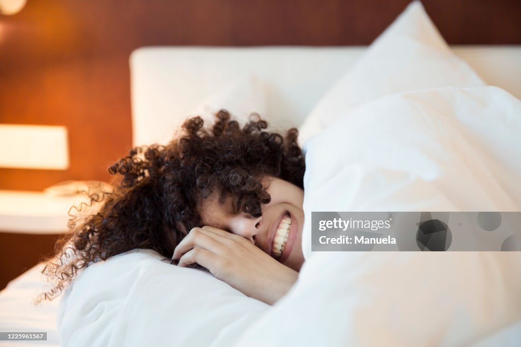 A smiling woman with dark curly hair in bed under the covers, head on a white pillow,.