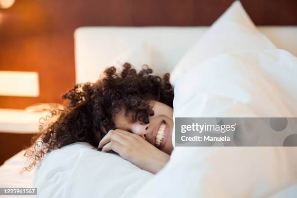 a smiling woman with dark curly hair in bed under the covers, head on a white pillow,. - bed woman stock pictures, royalty-free photos & images