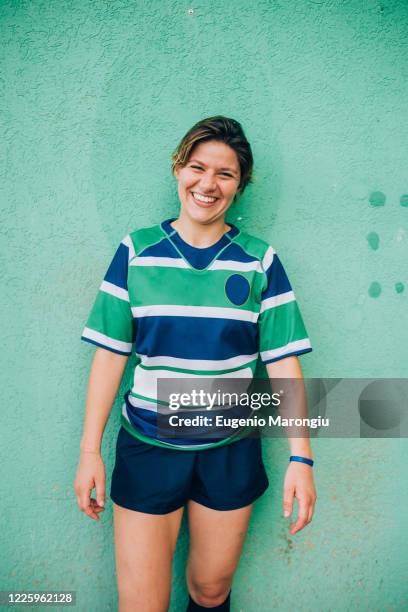 a woman wearing a blue, white and green rugby shirt standing against a green wall looking at the camera and smiling. - rugbyhemd stock-fotos und bilder