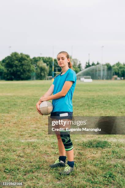 a woman standing on a training pitch in shorts and tee shirt with a knee strap holding a rugby ball. - rugby boot stock pictures, royalty-free photos & images