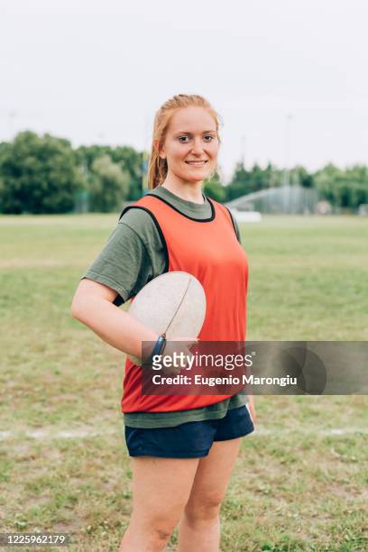 a woman standing on a training pitch in shorts and tee shirt holding a rugby ball. - rugby boot stock pictures, royalty-free photos & images