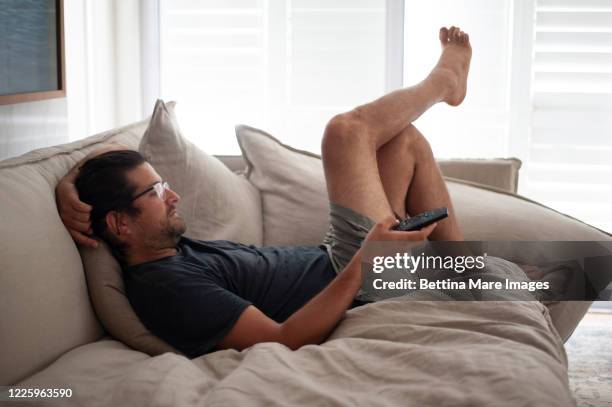 a man lounging on a sofa wearing shorts and a t shirt holding a remote control, staying at home. - boredom man stock pictures, royalty-free photos & images