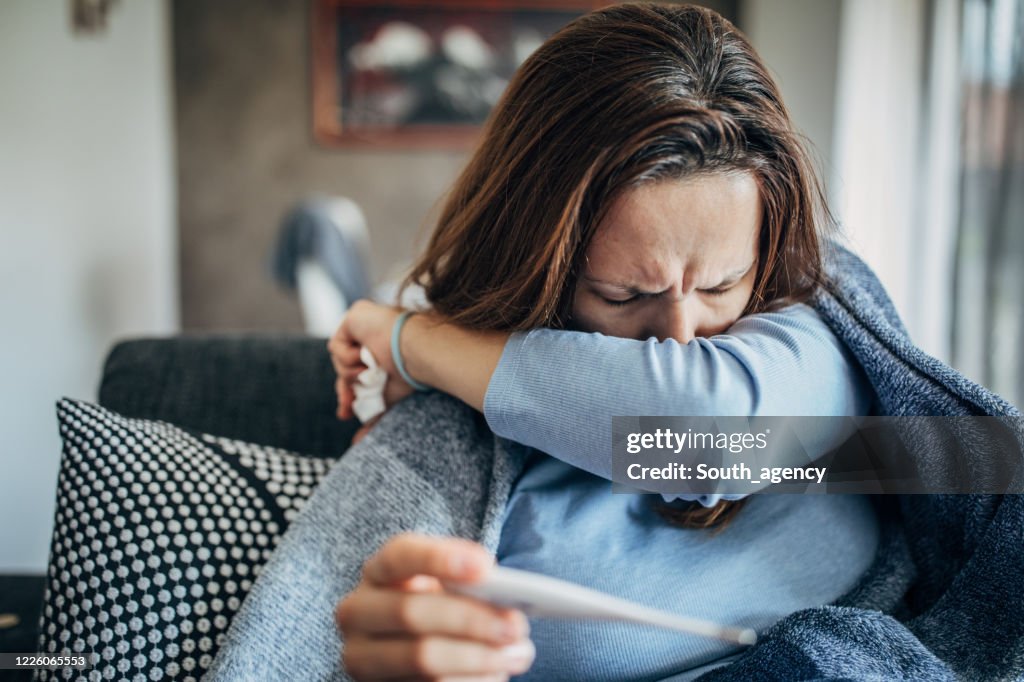 Frau mit Fiebersymptomen sitzt auf Sofa und hält Thermometer