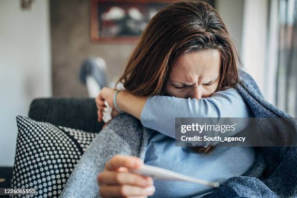 frau mit fiebersymptomen sitzt auf sofa und hält thermometer - stressed young woman sitting on couch stock-fotos und bilder