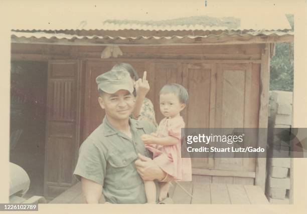 Mid-shot of a young serviceman, standing in front of a wooden building on a sunny day, smiling at the camera and holding a small girl in one arm...