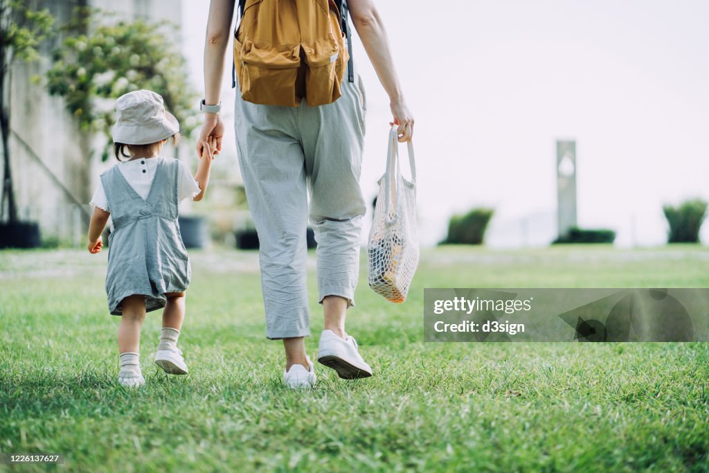 Cropped shot of young Asian mother carrying groceries with cotton mesh eco bag. Walking hand in hand with little daughter across parkland after grocery shopping together. Zero waste concept