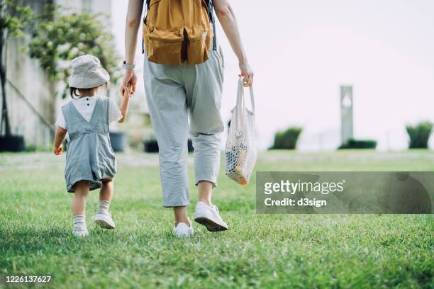 cropped shot of young asian mother carrying groceries with cotton mesh eco bag. walking hand in hand with little daughter across parkland after grocery shopping together. zero waste concept - asian family shopping foto e immagini stock