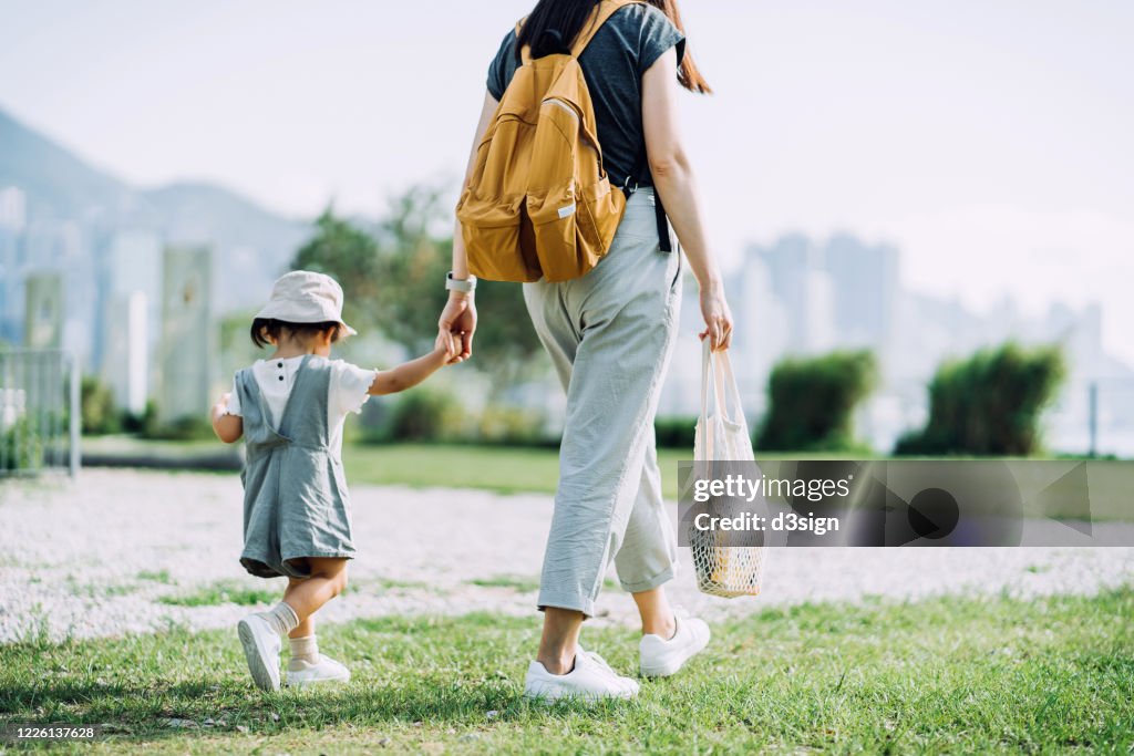 Cropped shot of young Asian mother carrying groceries with cotton mesh eco bag. Walking hand in hand with little daughter across parkland after grocery shopping together. Zero waste concept