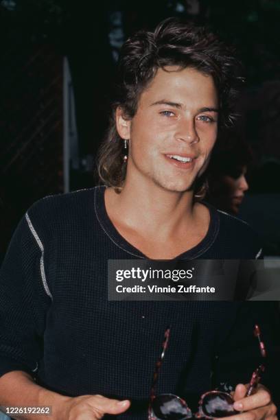 American actor Rob Lowe wearing a dark crew neck top with white trim, and holding a pair of sunglasses with tortoiseshell frames, circa 1985.