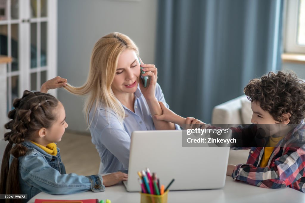 Blonde female talking on mobile and working on laptop, distracted by curly boy and dark-haired girl