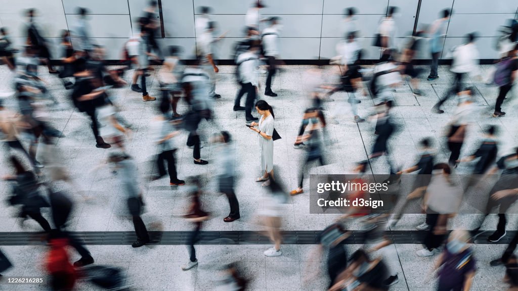 Young Asian woman using smartphone surrounded by commuters rushing by in subway station during office peak hours in the city