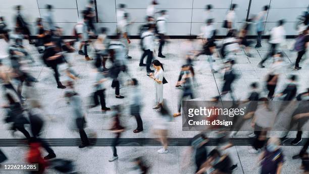 young asian woman using smartphone surrounded by commuters rushing by in subway station during office peak hours in the city - beengt stock-fotos und bilder