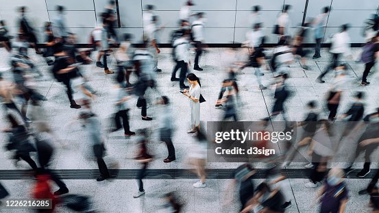 Young Asian woman using smartphone surrounded by commuters rushing by in subway station during office peak hours in the city Young Asian woman using smartphone surrounded by commuters rushing by in subway station during office peak hours in the city