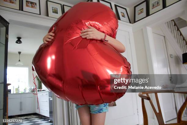 little boy standing in his living room holding up a big red heart shaped balloon in front of himself. - embracing heart stock pictures, royalty-free photos & images