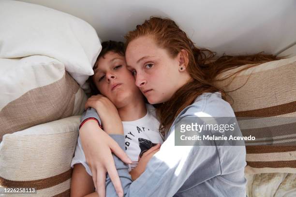Teenage girl and her little brother cuddled together on a daybed in their home, looking very sleepy.