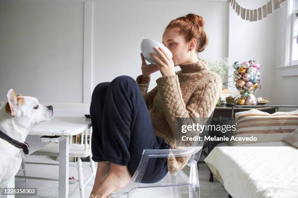 teenage girl sitting at her dining room table curled up in a modern chair and sipping out of a large white bowl. her dog is halfway in the frame watching her. profile shot. looking up while sipping - love photos et images de collection