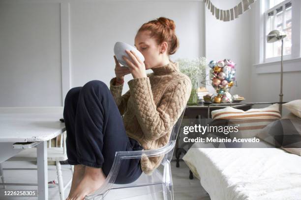 teenage girl sitting at her dining room table curled up in a modern chair sipping out of a large white bowl. her dog is halfway in the frame watching her. profile shot. - love photos et images de collection