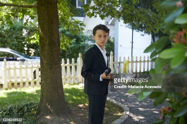 young boy with brown curly hair brown eyes standing in the front yard standing next to tree wearing a suit with white formal shirt underneath navy blue suit jacket small white flower in his jacket - brown suit 個照片及圖片檔