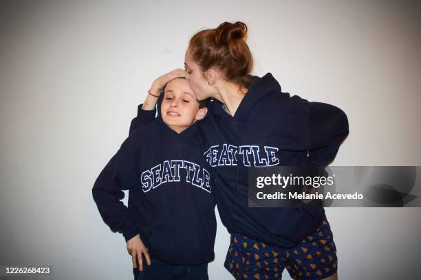 young boy with brown curly hair brown eyes with his older sister who has red hair both are standing in front of a white wall wearing matching sweatshirts posing for the camera camera flash older sister is kissing brothers head - teasing stock pictures, royalty-free photos & images
