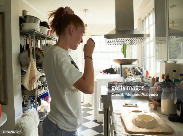 teenage girl with red long straight hair brown eyes and freckles standing up in the kitchen making homemade pasta with flour and raw egg cracked in the middle on a cutting board hair putting up in a pony tail wearing a t shirt looking down at the counter - skinny teen ストックフォトと画像