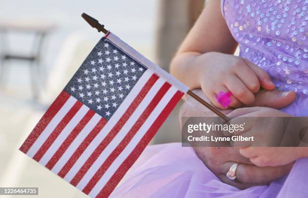 a little girl, in in a purple sparkly dress, says the pledge of allegiance while holding an american flag while sitting on her dad’s lap - s allegiance season 1 stock-fotos und bilder