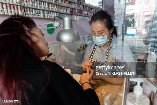 Nail technician wearing a face mask or covering due to the COVID-19 pandemic, sits behind a perspex safety screen as she paints the nails of a...