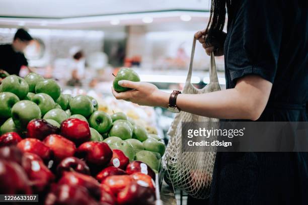 cropped shot of young asian woman shopping for fresh organic groceries in supermarket. she is shopping with a cotton mesh eco bag and carries a variety of fruits and vegetables. zero waste concept - frische stock-fotos und bilder