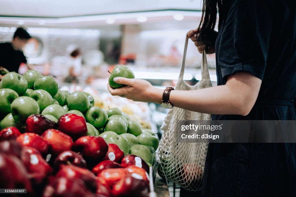 Cropped shot of young Asian woman shopping for fresh organic groceries in supermarket. She is shopping with a cotton mesh eco bag and carries a variety of fruits and vegetables. Zero waste concept