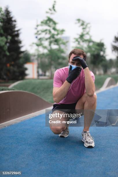 Sportsman with medical mask and gloves, smartphone and earbuds working out, jogging in urban surroundings. Exhausted man from jogging and medical mask usage. Trouble with breathing.