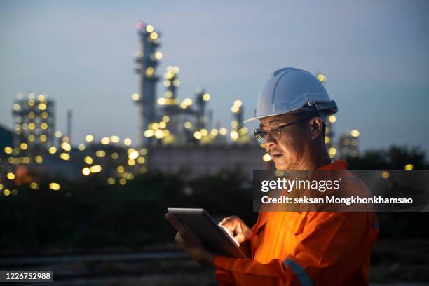 asian man engineer using digital tablet working late night shift at petroleum oil refinery in industrial estate. chemical engineering, fuel and power generation, petrochemical factory industry concept - industrial estate ストックフォトと画像