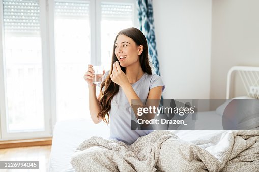 Smiling healthy young woman taking supplements and drinking water in bed Smiling healthy young woman taking supplements and drinking water in bed