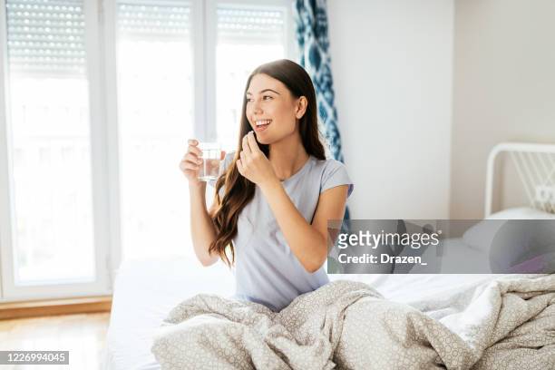 smiling healthy young woman taking supplements and drinking water in bed - moving activity stock pictures, royalty-free photos & images