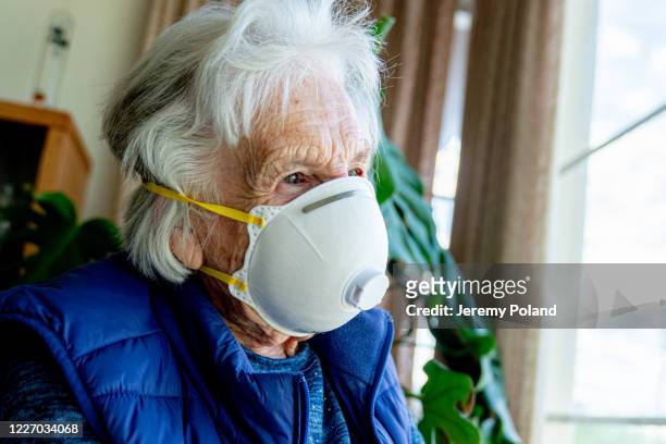close-up shot of distraught elderly senior caucasian woman looking out the window feeling loneliness wearing an n95 protective face mask to prevent the spread of covid sars ncov 19 coronavirus swine flu h7n9 influenza illness during cold and flu season - n95 respirator mask stock-fotos und bilder